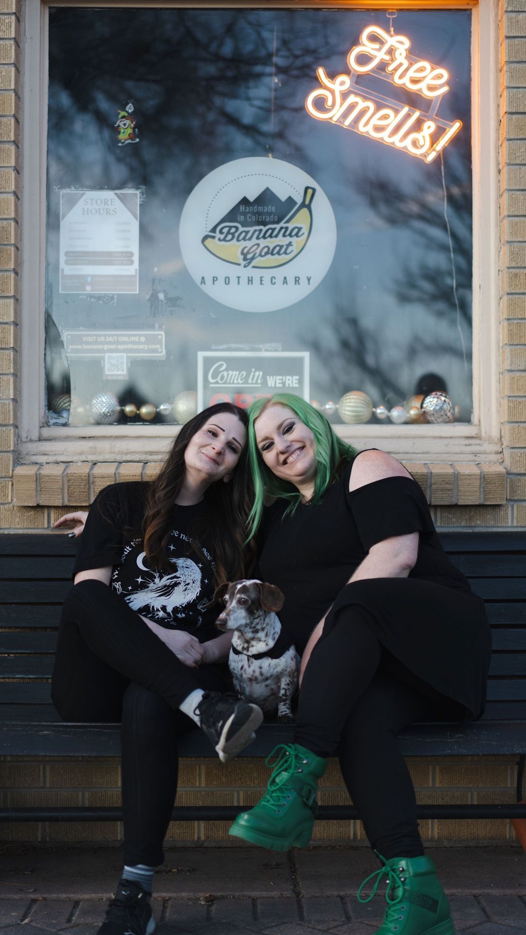 Two women are sitting on a bench with a dog in front of a store window.