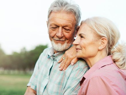 Two older adults smiling outdoors, leaning close together in a green park