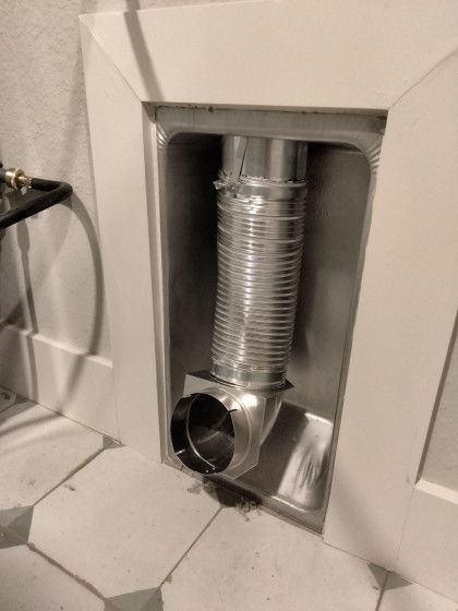 Flexible silver dryer vent hose in a recessed wall opening above a tiled floor.
