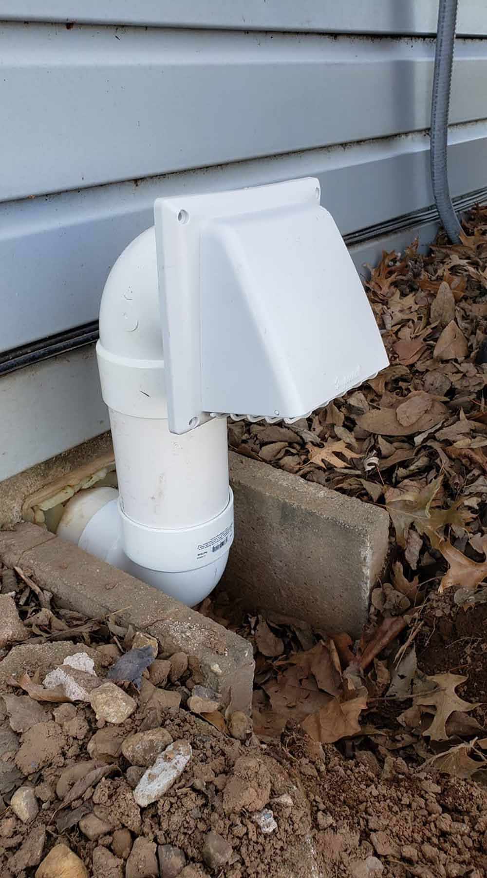 White vent cap on a PVC pipe beside a house foundation, surrounded by dirt and dry leaves.