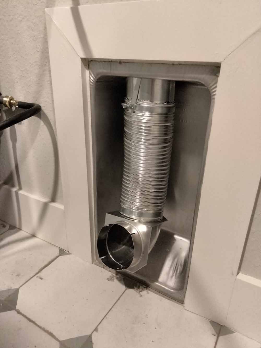 Flexible silver vent duct inside a recessed wall opening above a tiled floor.