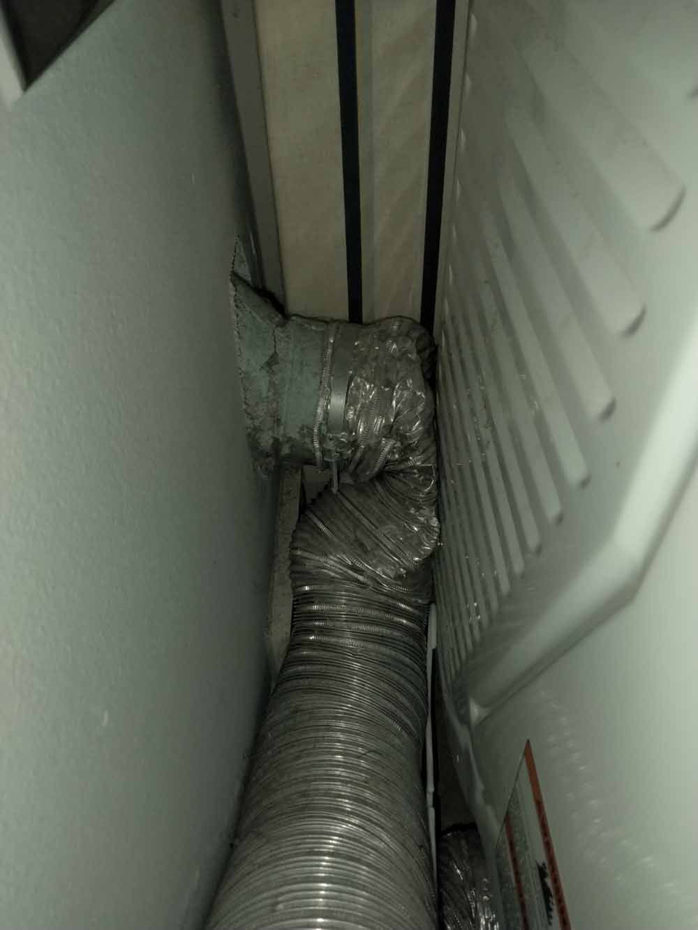 Silver flexible duct running between two white appliances in a narrow gap