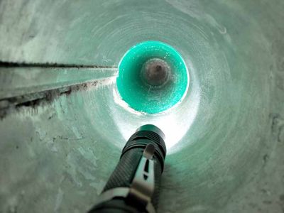Looking up inside a green-tinted pipe toward a circular opening, with a drill or tool in the foreground