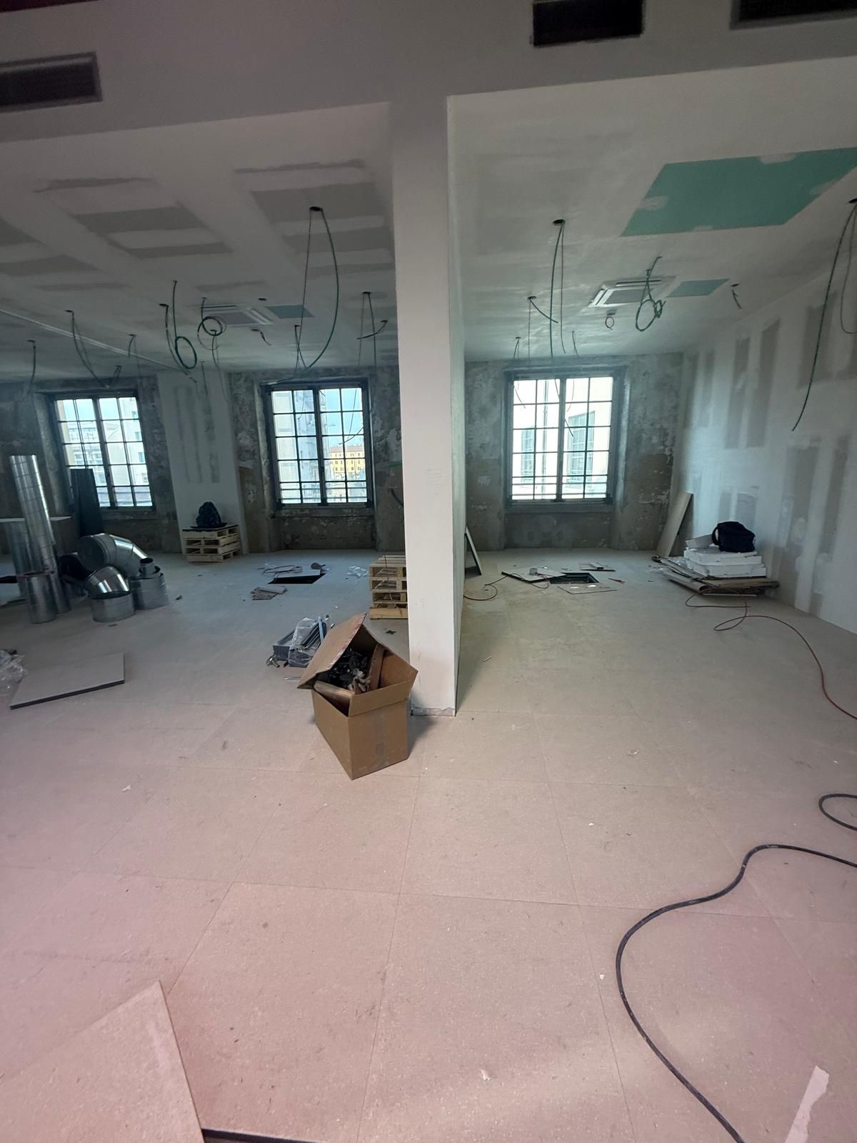 Cantiere di ristrutturazione in un ampio locale con cavi a vista, finestre, detriti e una colonna portante al centro.