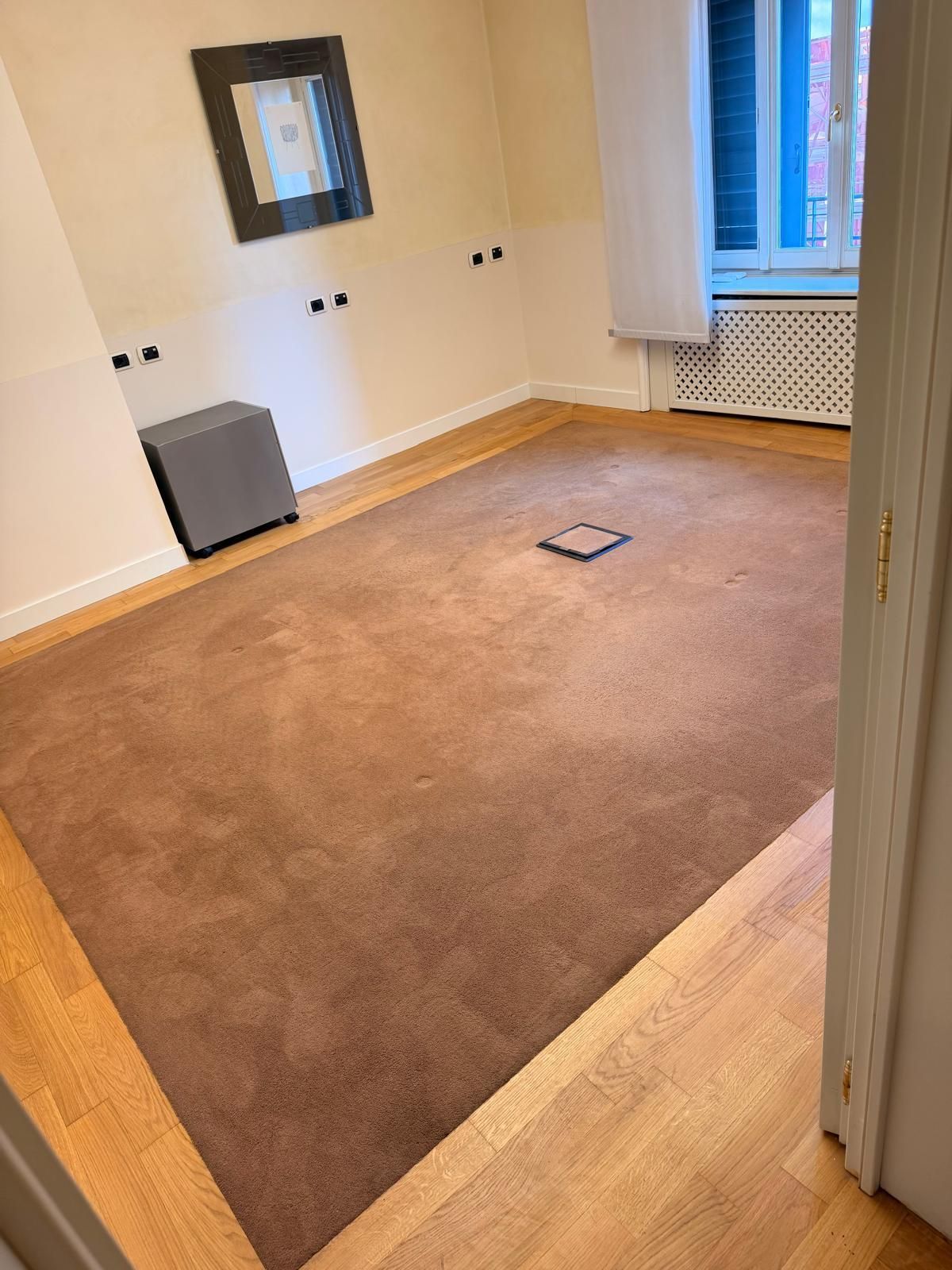Stanza vuota e non arredata, con moquette beige, pareti bianche e un piccolo specchio a parete vicino a una finestra.