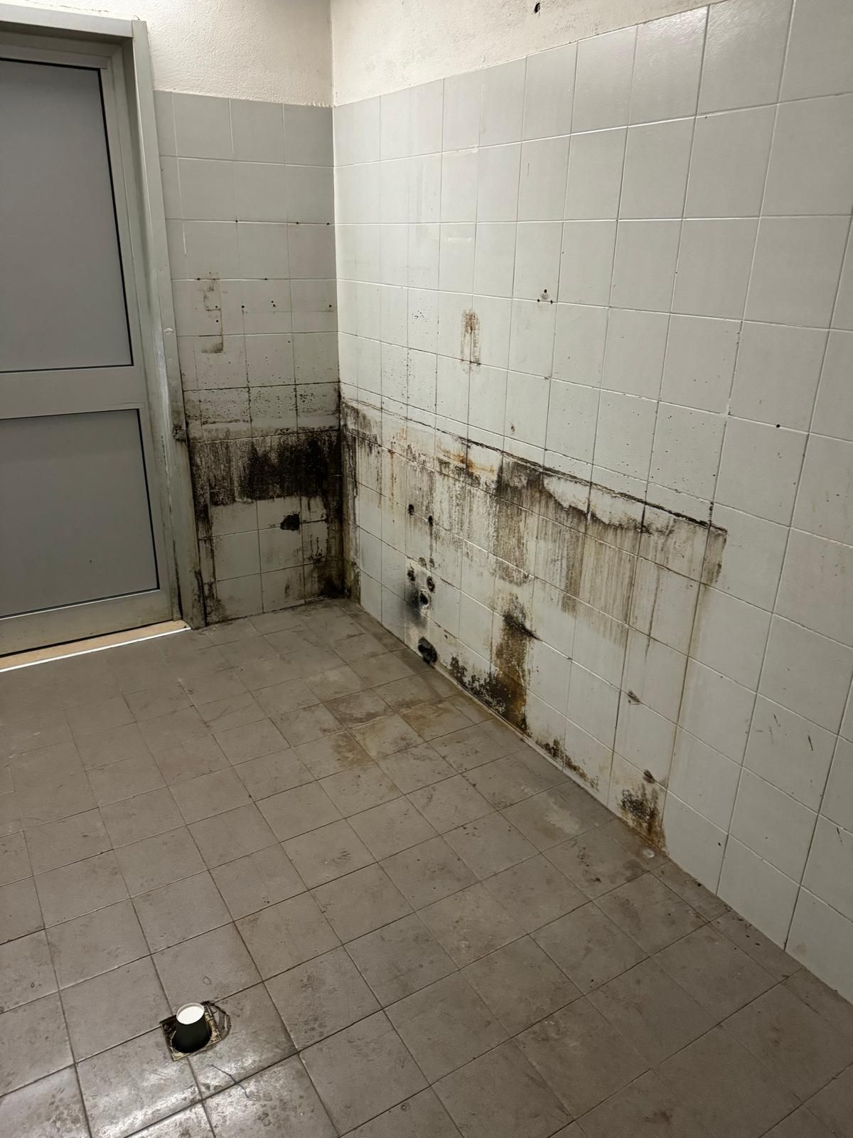 Bagno pubblico piastrellato sporco, con macchie di muffa sulla parete e uno scarico a pavimento vicino a una porta grigia