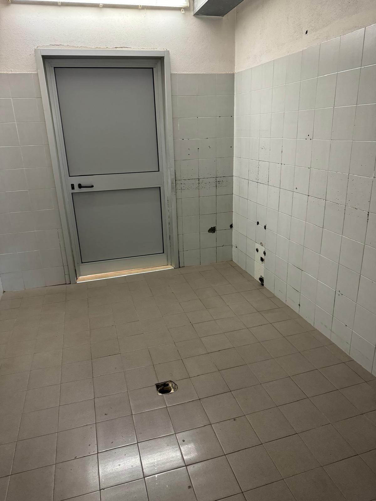 Piccola stanza piastrellata con una porta grigia chiusa e uno scarico a pavimento al centro.