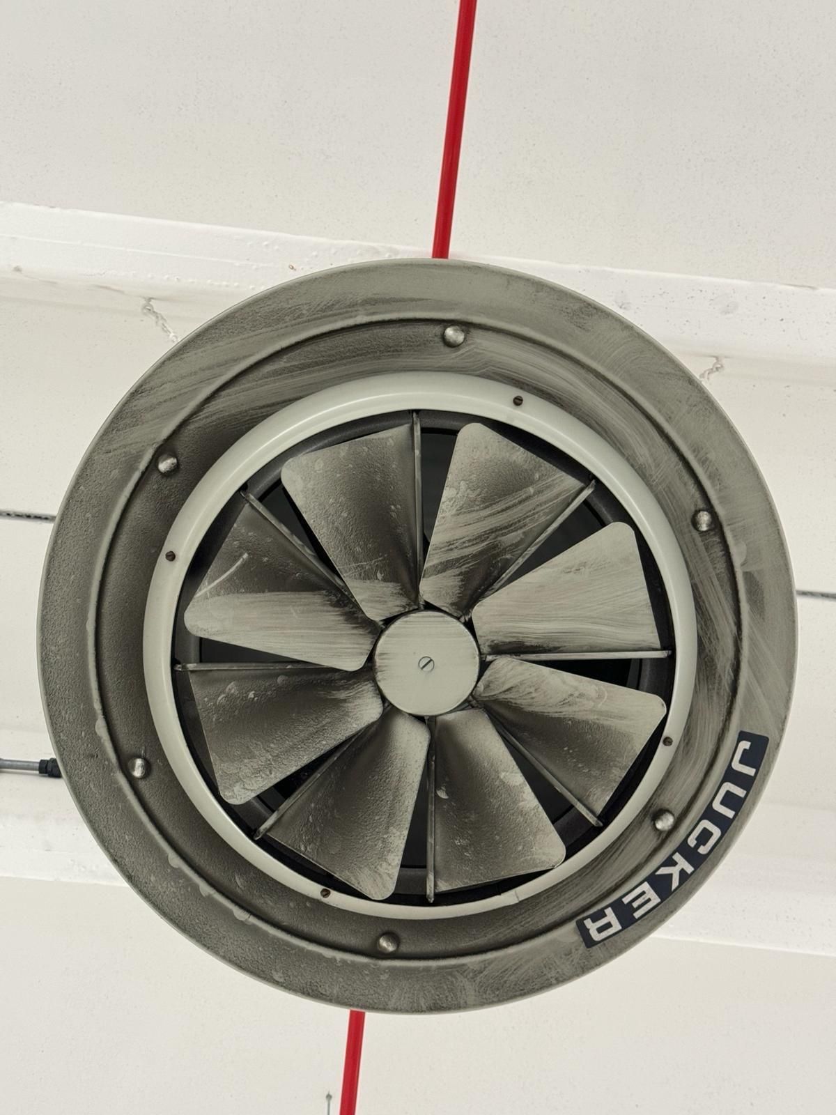 Ventilatore industriale rotondo con pale grigie e cavo rosso, montato su una parete bianca.