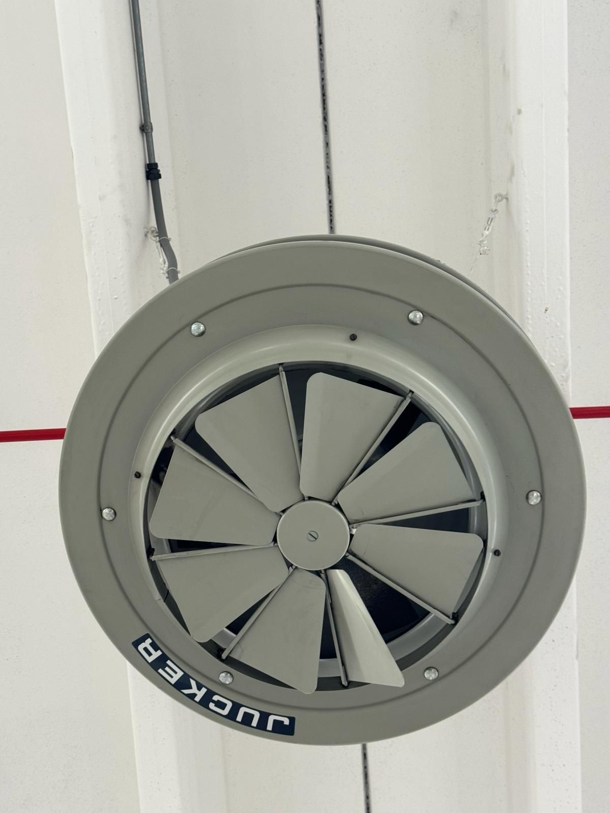 Ventilatore da soffitto in un alloggiamento di metallo grigio, contro una parete bianca.