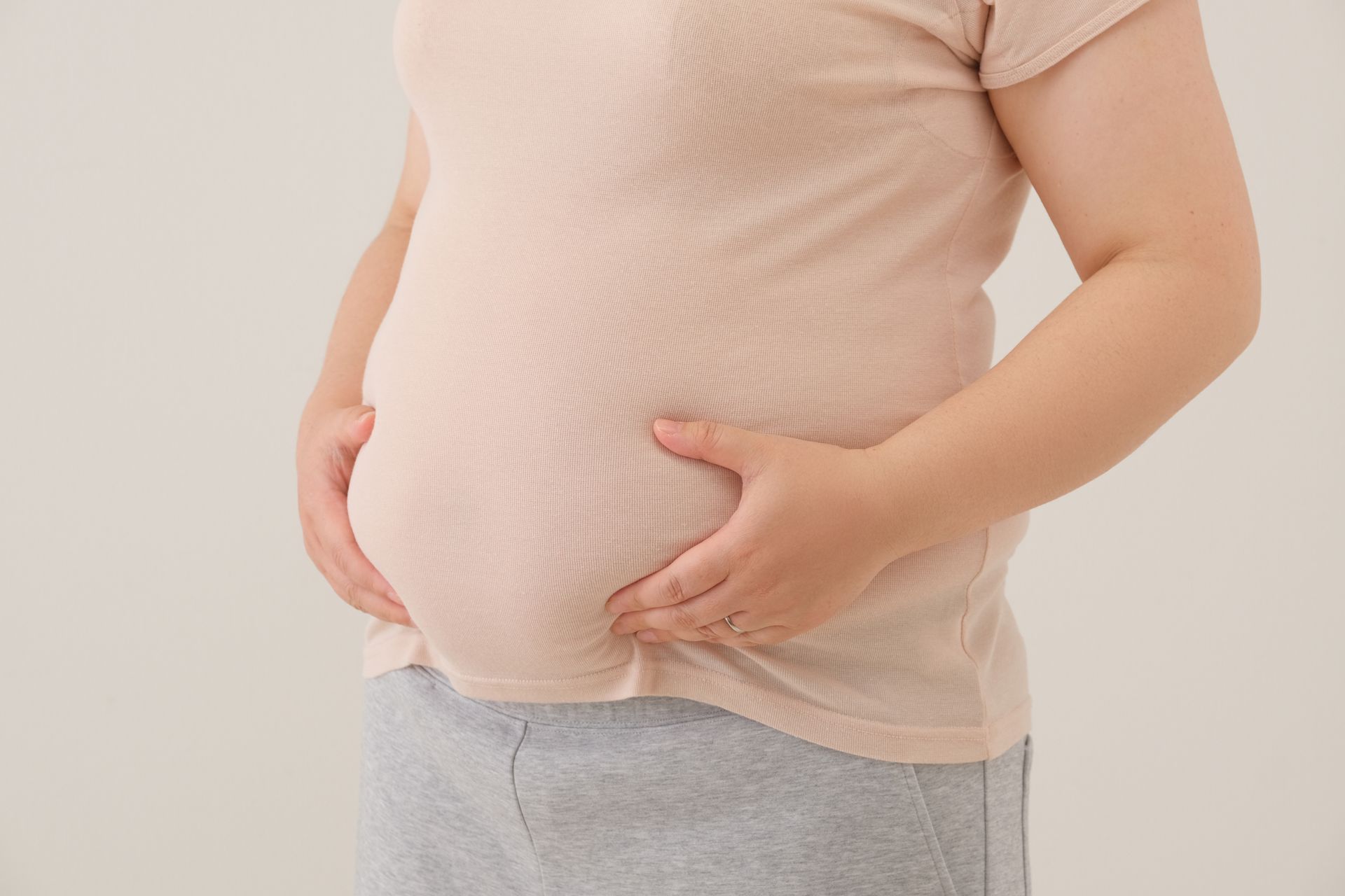 Person holding a large pregnant belly in a light pink shirt and gray pants Person holding a large pregnant belly in a light pink shirt and gray pants