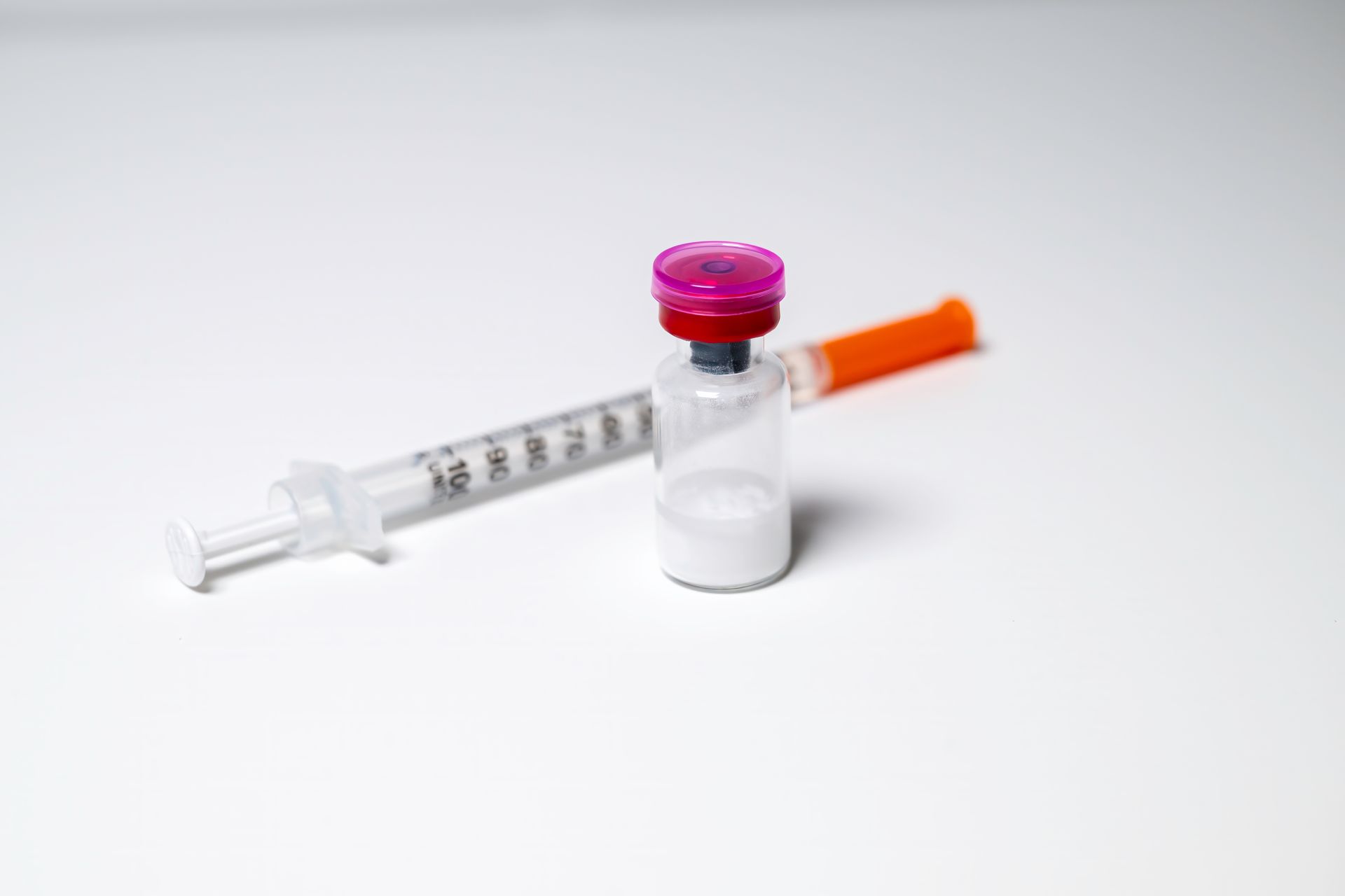 Syringe beside a small medicine vial with a pink cap and orange needle cover on a white background