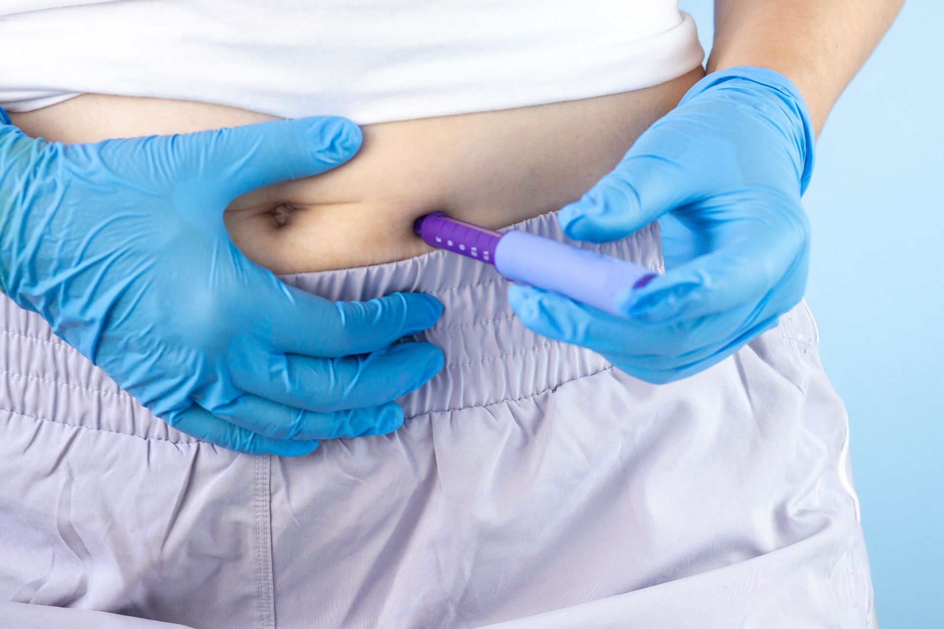 Gloved hands inject medication into a person’s abdomen with a purple syringe Gloved hands inject medication into a person’s abdomen with a purple syringe