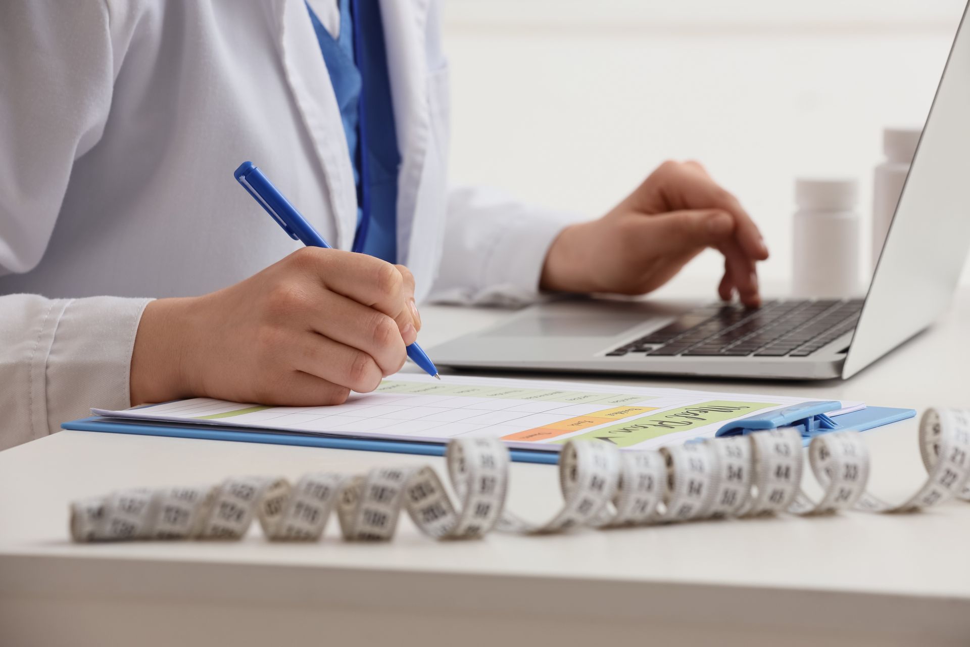 Medical provider documenting a patient's weight loss treatment plan with a laptop, medication, and tape measure. Medical provider documenting a patient's weight loss treatment plan with a laptop, medication, and tape measure.