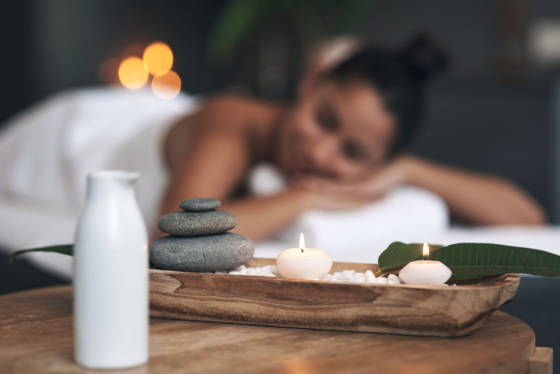 Spa scene with candles, stacked stones, a white bottle, and a blurred person resting in the background Spa scene with candles, stacked stones, a white bottle, and a blurred person resting in the background