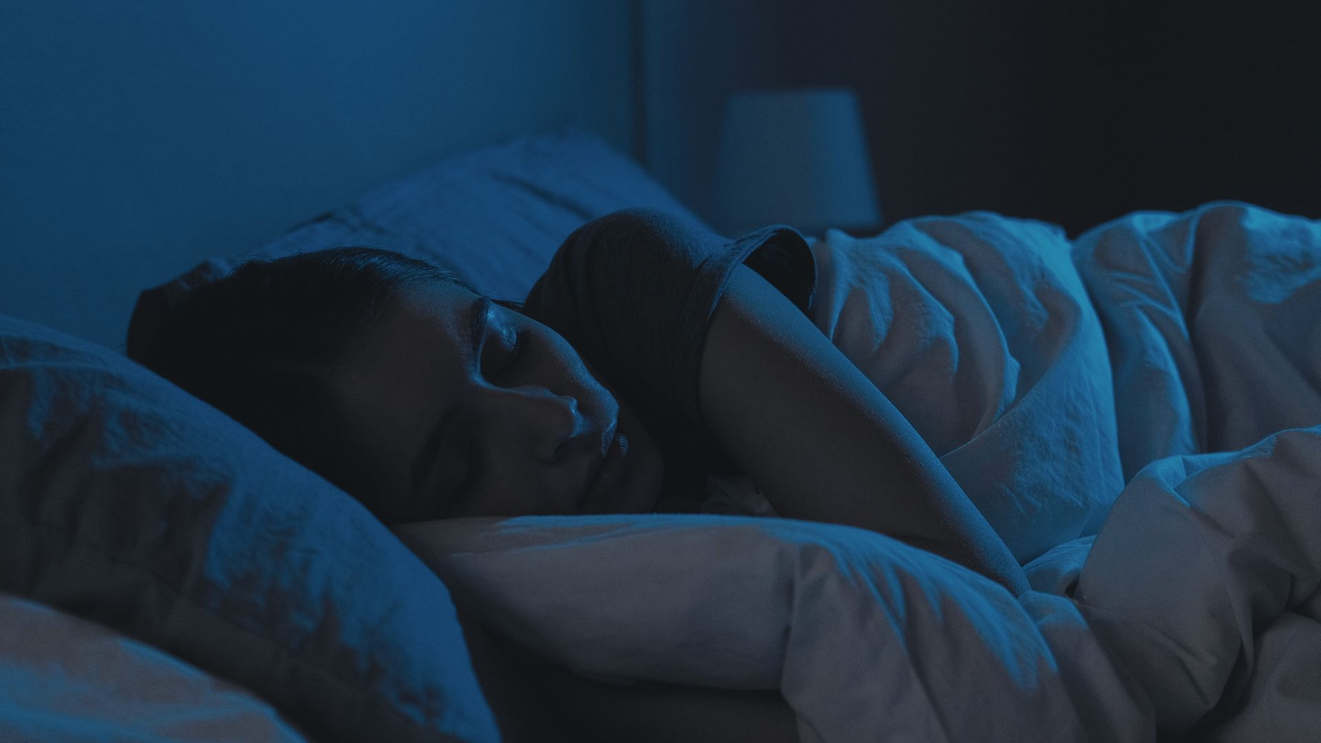 Person sleeping in bed under white blanket in dim blue light Person sleeping in bed under white blanket in dim blue light