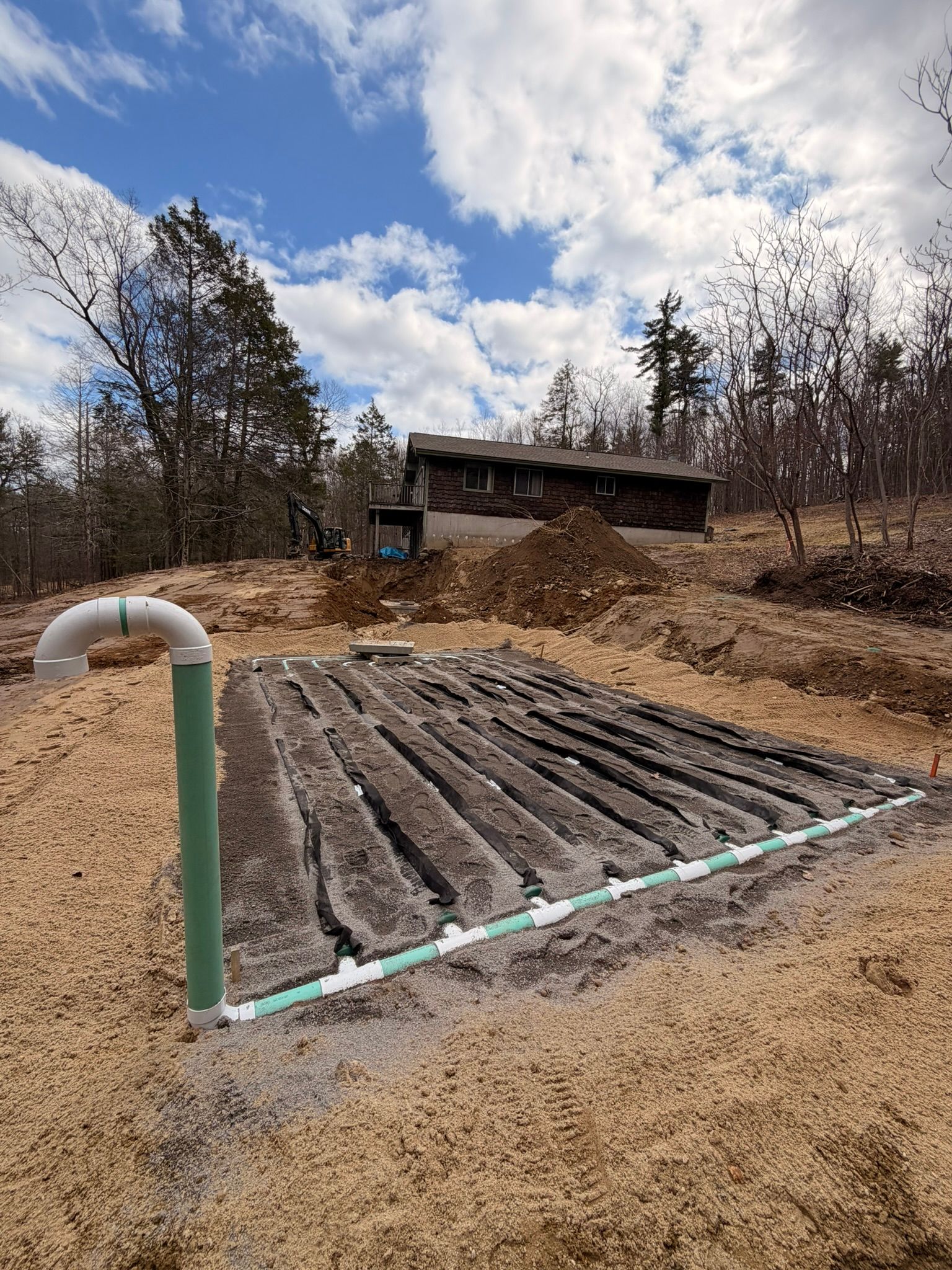 Raised garden bed with dark soil rows, a white pipe, and a house under cloudy sky in a rural yard