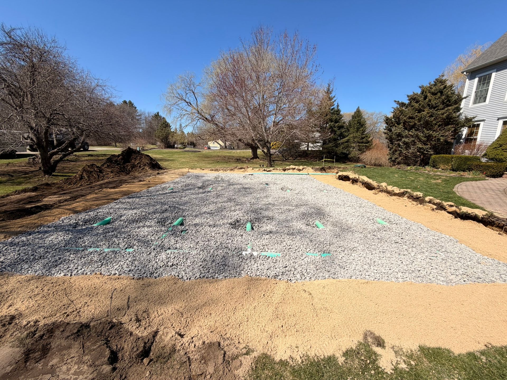 Gravel-covered driveway under construction beside a suburban home, with trees and blue sky in the background