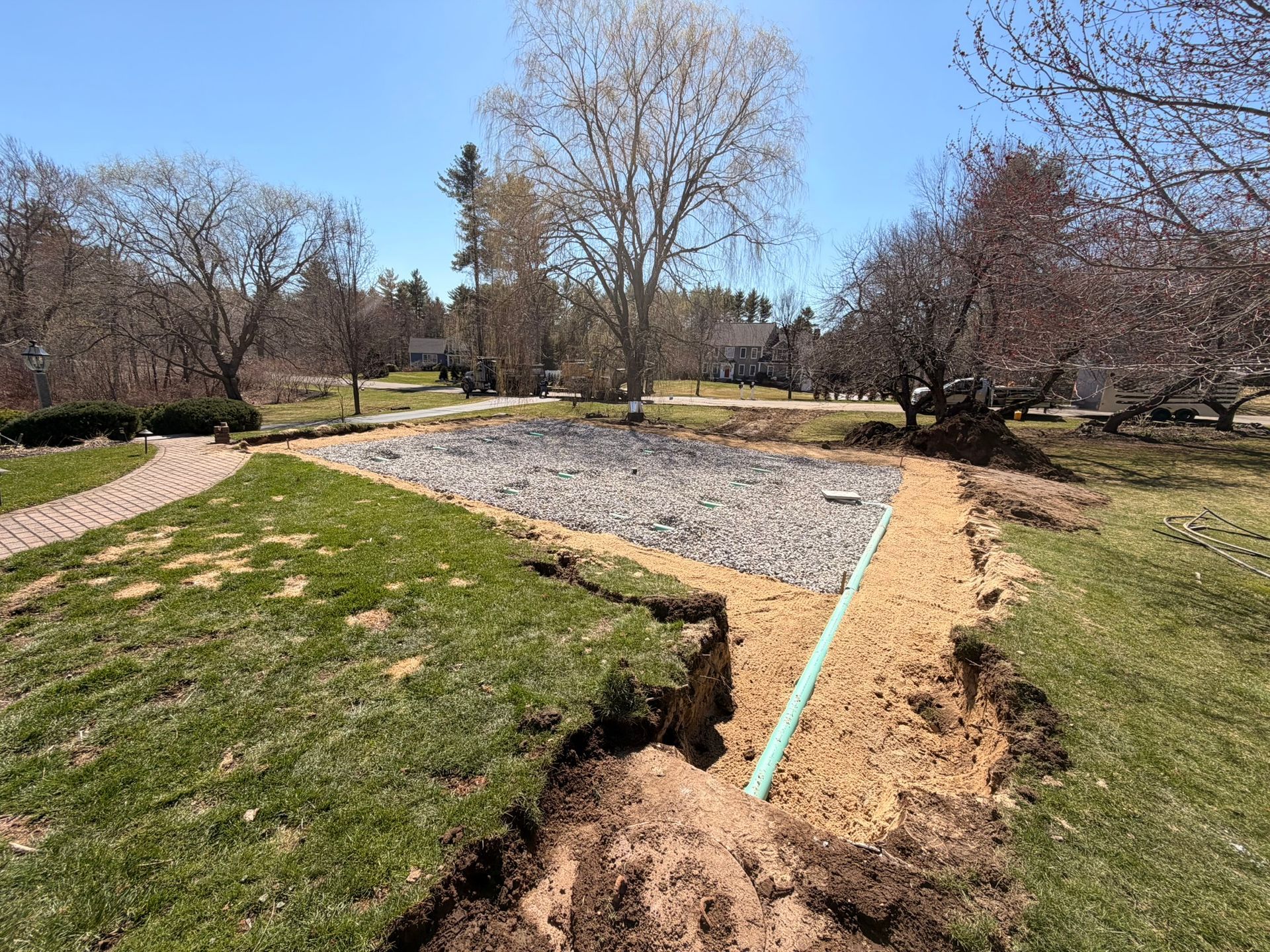 Park landscape with bare trees, gravel path, and a construction trench with a blue pipe and exposed soil.