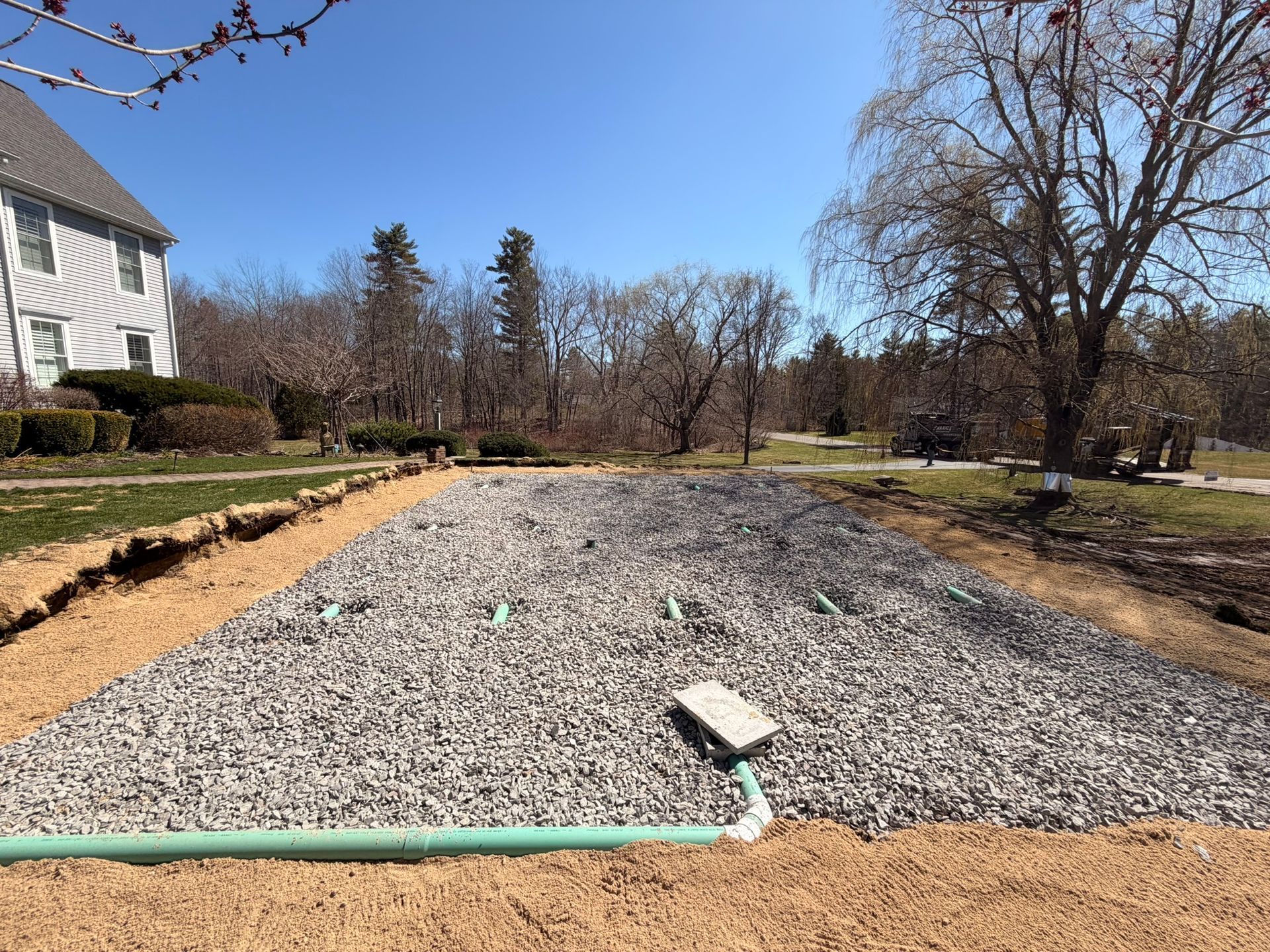 Gravel-filled drainage trench beside a house, with a green hose in the foreground and trees lining the yard