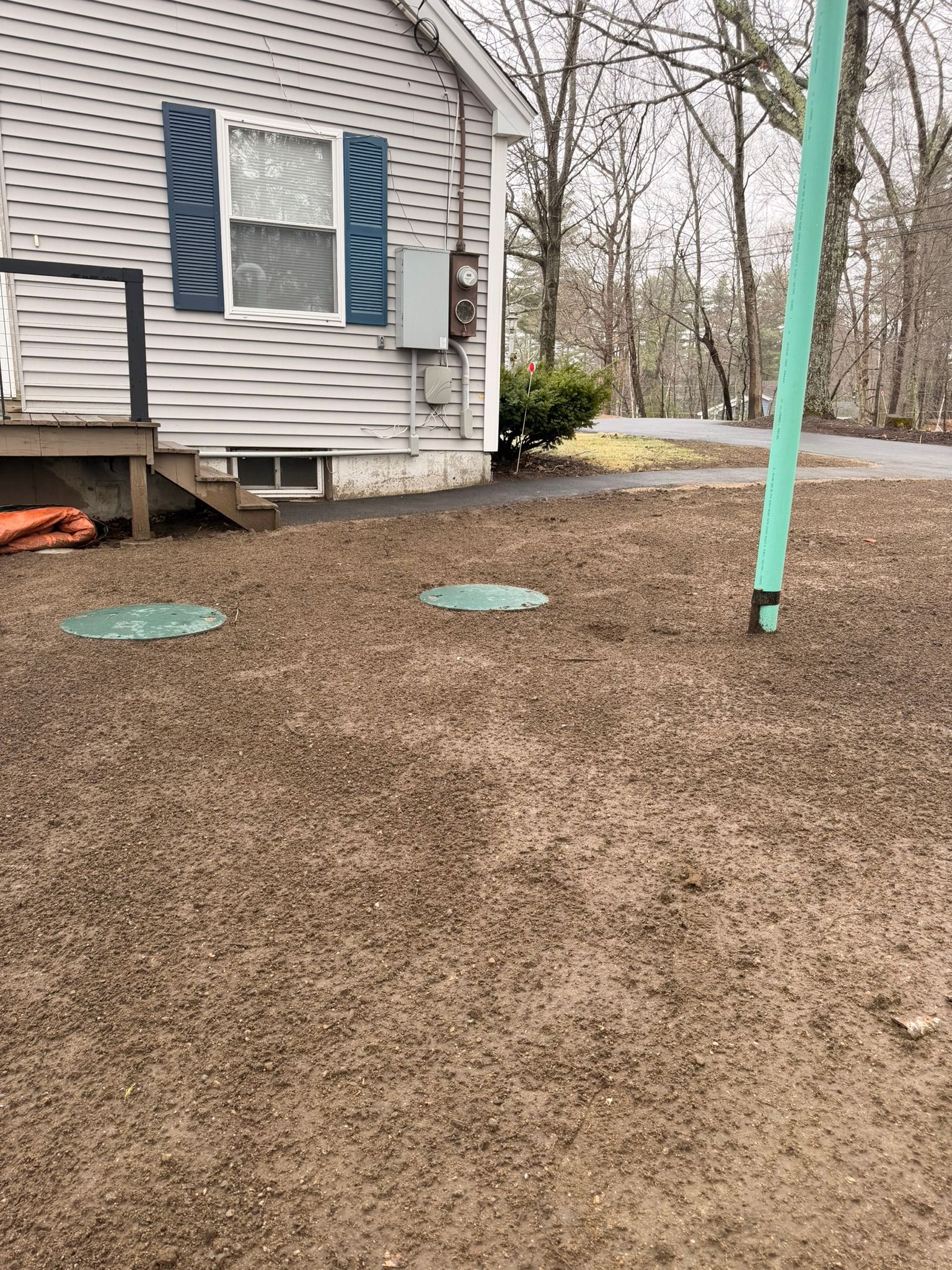 Backyard beside a gray house with blue shutters, gravel ground, and a teal pole in the foreground