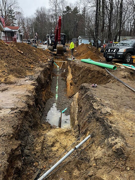 Construction site: excavator in a deep trench with pipes, dirt piles, and a worker in a yellow vest.