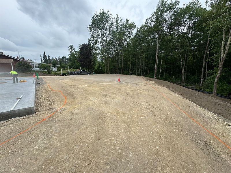 Construction site with graded dirt lot, concrete pad, trees, and cloudy sky.