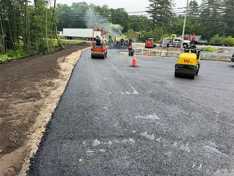 Road paving in progress; asphalt spread by crew with rollers. Grey pavement, orange cones, construction vehicles.