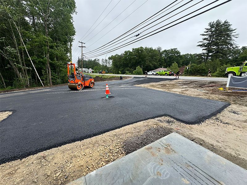 Asphalt paving in progress on a road, with machinery, a traffic cone, and utility poles visible.