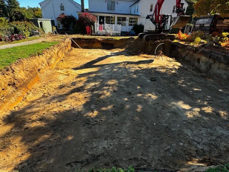 Large excavated area in front of a white house; excavator visible, sunny day.