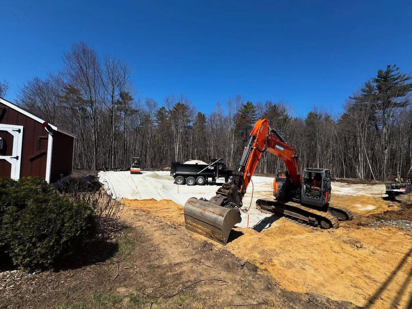 An excavator digging in a dirt lot next to a red barn. A dump truck and other equipment are visible. Bright blue sky.
