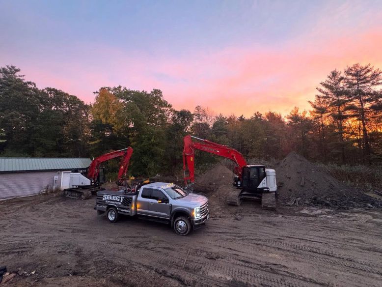 A silver flatbed truck with two excavators on a construction site, under a colorful sunset.