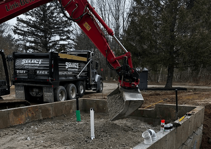 A red excavator dumping gravel into a concrete foundation next to a black dump truck in an outdoor setting.