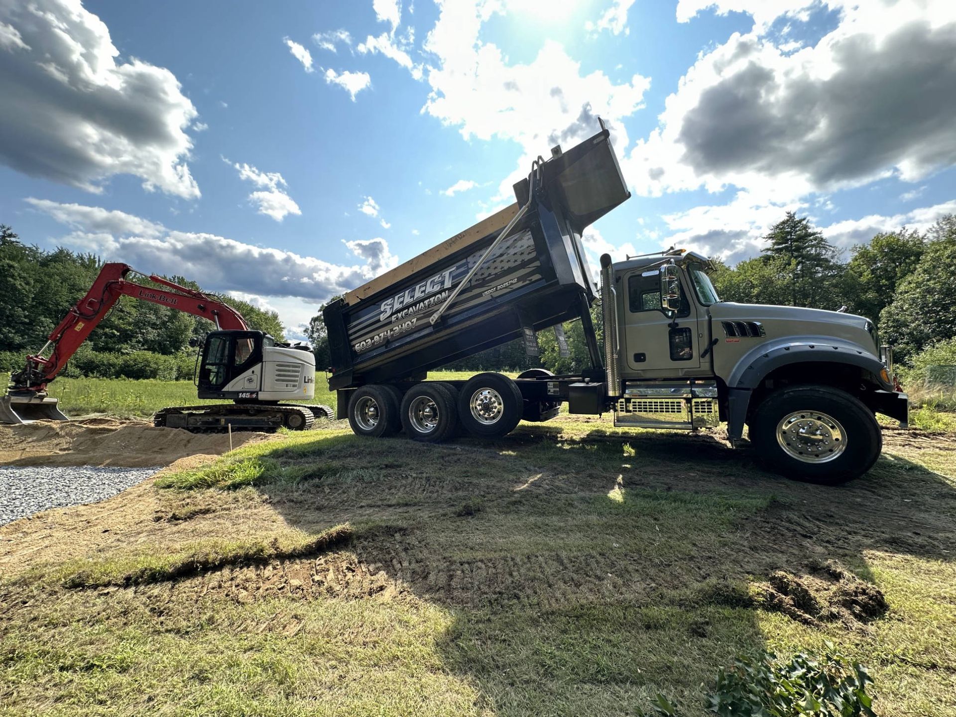 Excavator filling a dump truck with gravel on a grassy site under a cloudy sky.