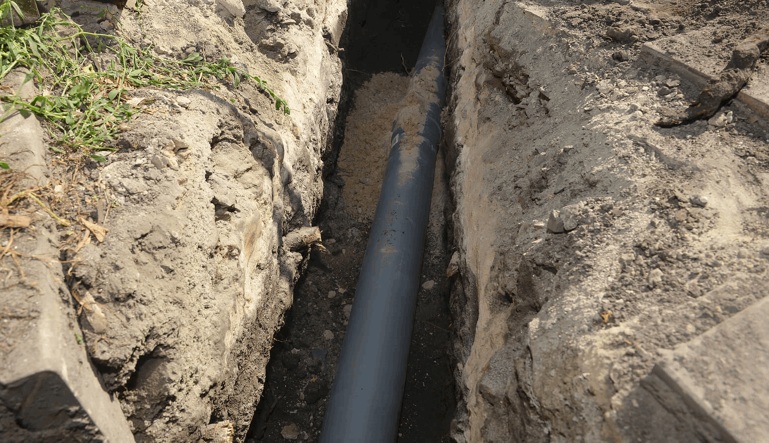 A gray pipe laid in a narrow, freshly dug trench, with dirt and gravel along the pipe.