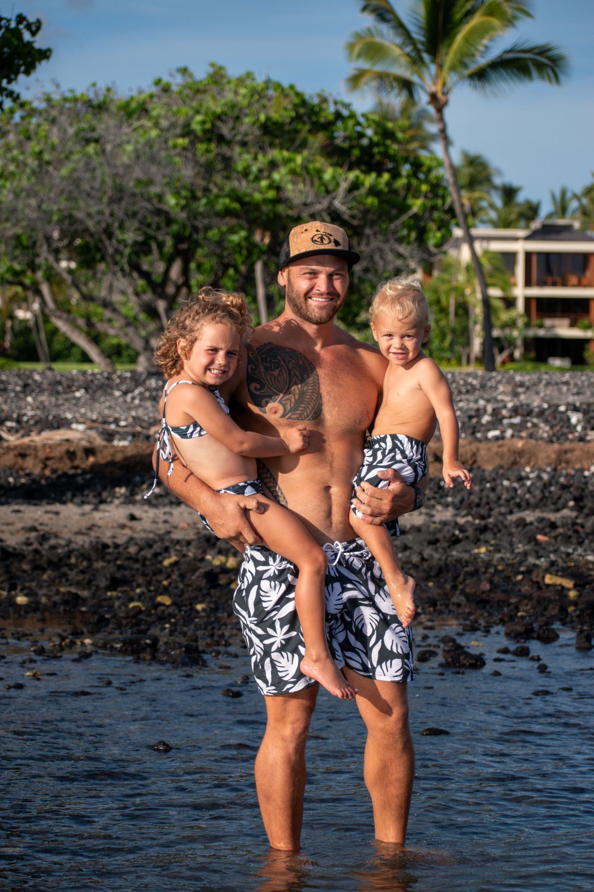 Man standing in shallow water holding two children, with palm trees and a beach resort in the background
