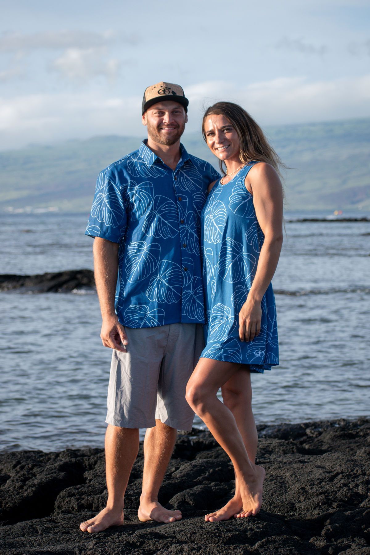 Two people standing on rocks by the ocean, wearing matching blue outfits, smiling at the camera.