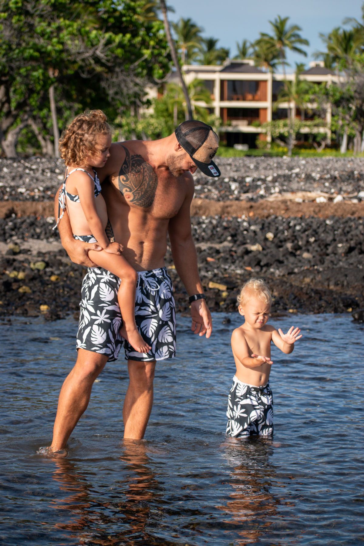 Family standing in shallow ocean water near a rocky shore, with palm trees and buildings in the background.