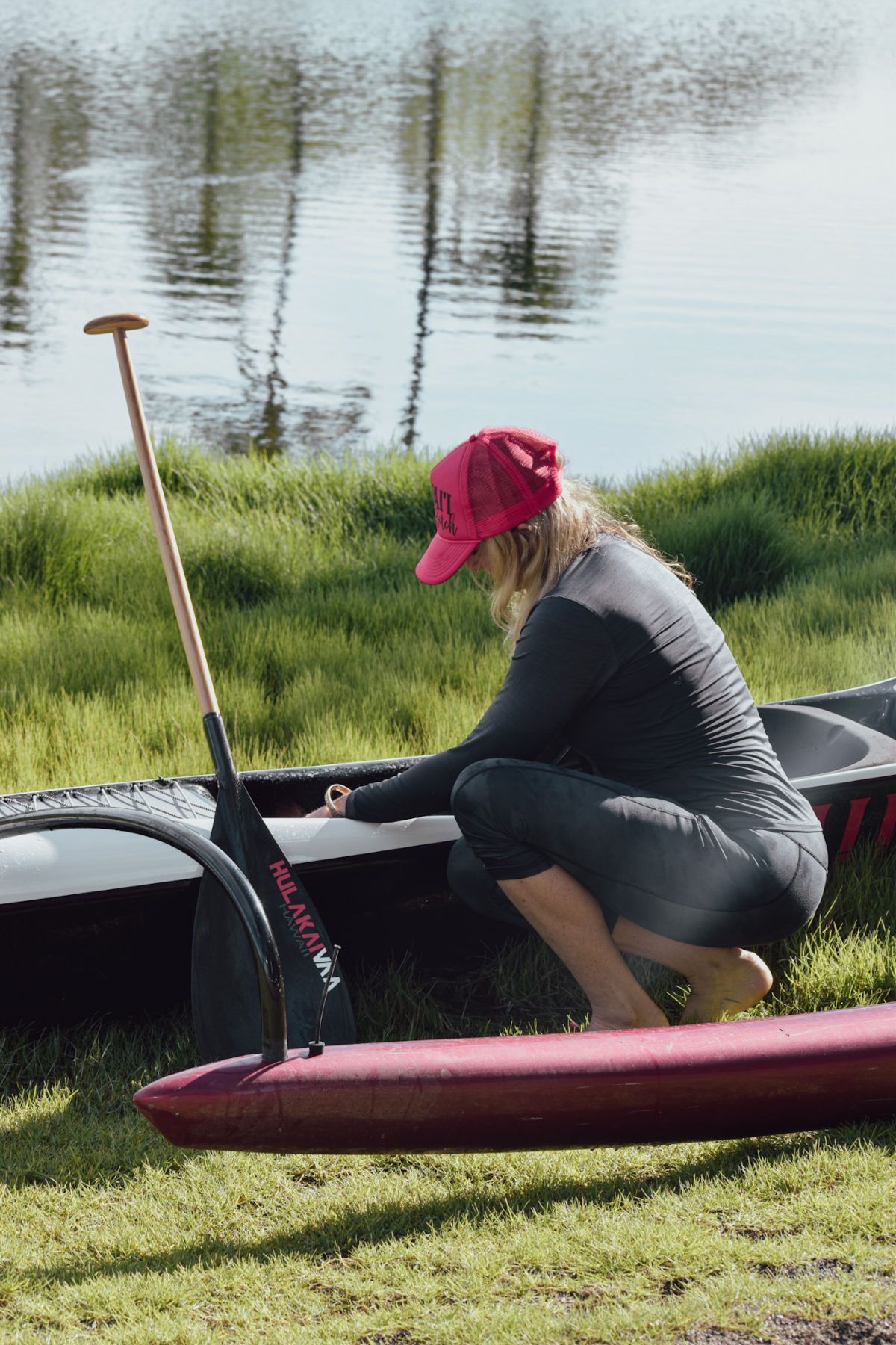 Person in a red cap kneels beside a black canoe by a calm lakeshore, holding a paddle.