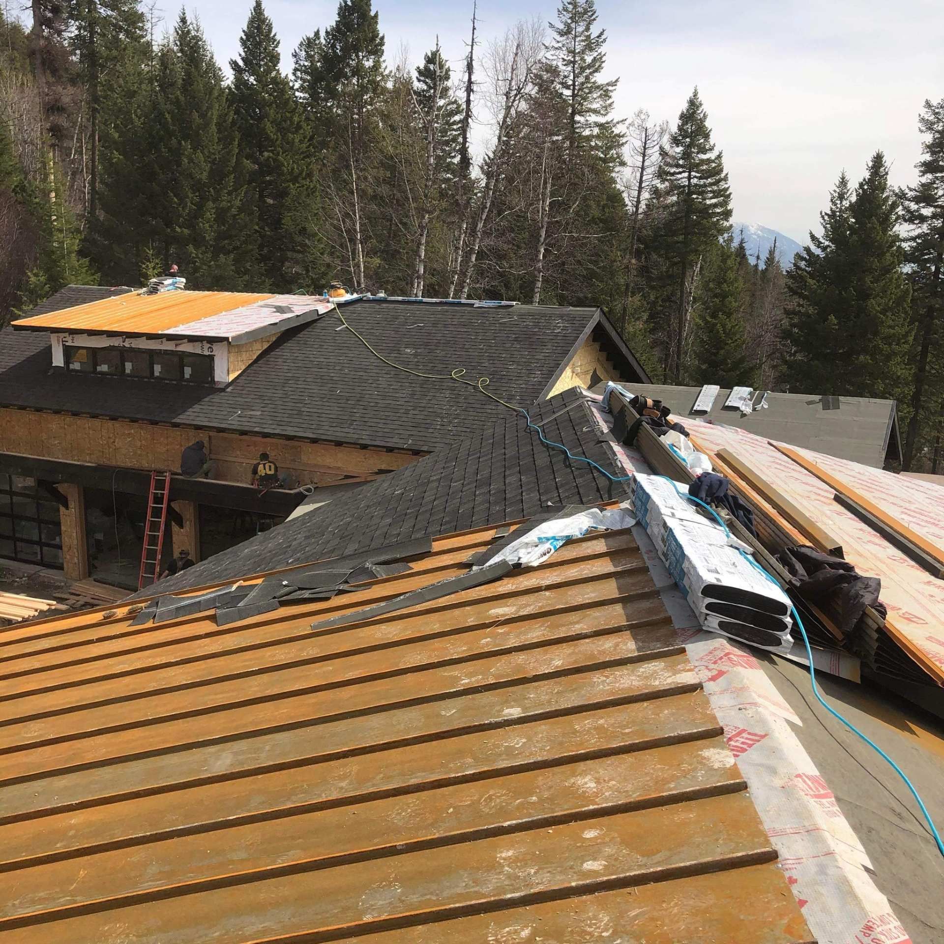 Partially reroofed house with exposed underlayment and stacks of roofing materials amid tall trees