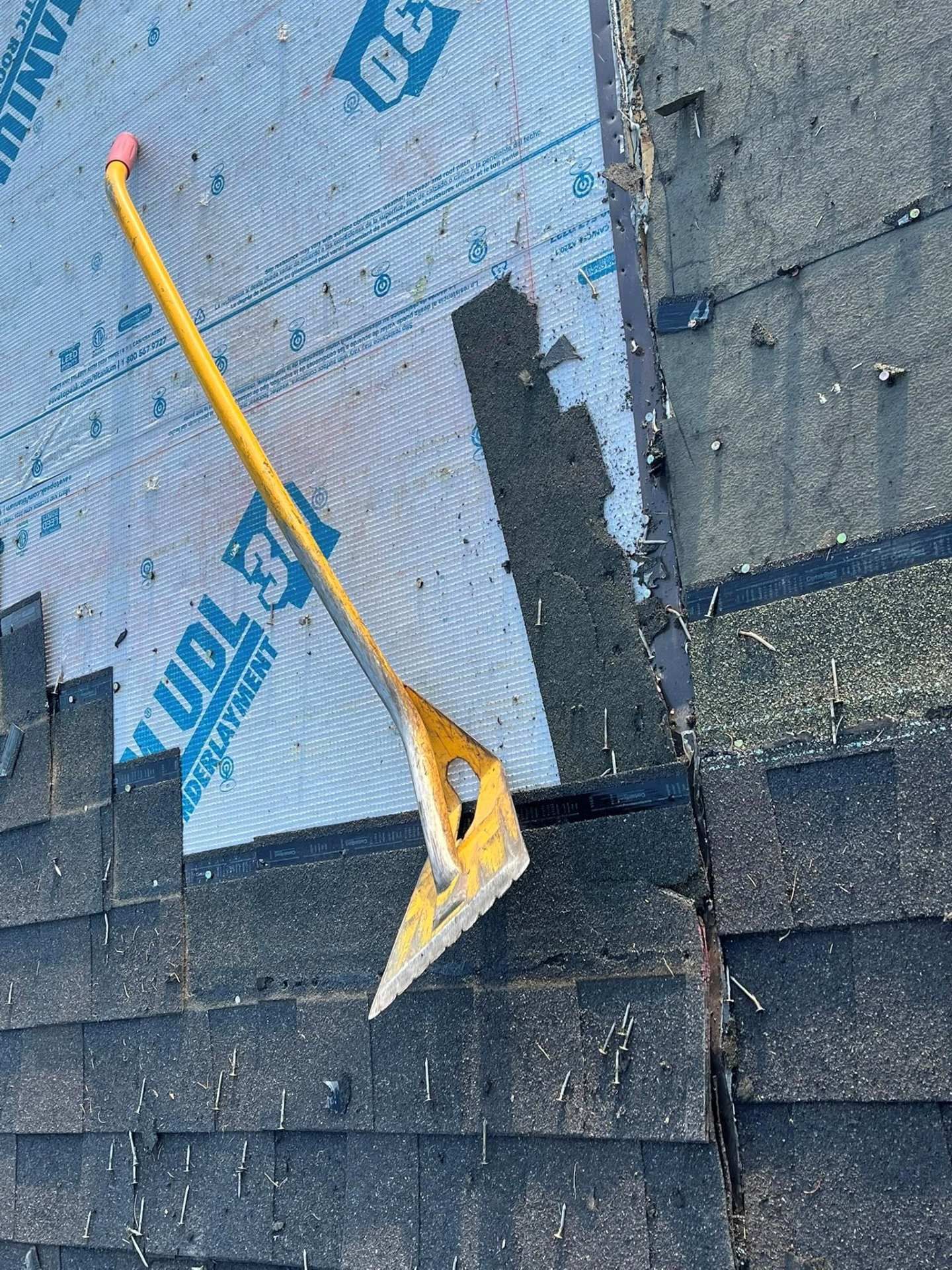 Yellow roof rake resting on roof shingles beside blue underlayment and exposed roofing area