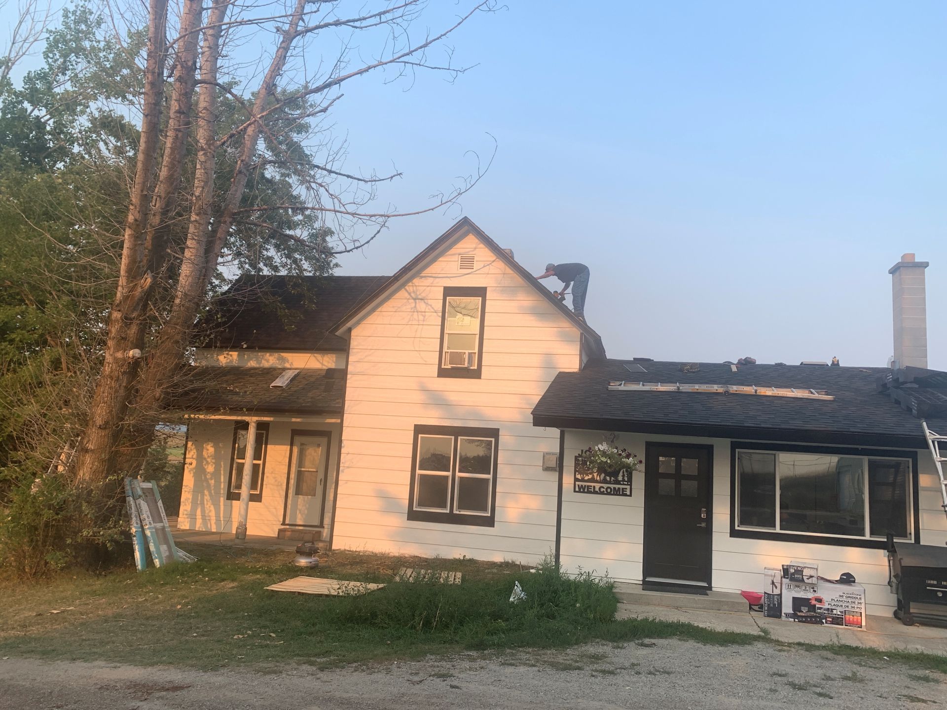 Small white house with dark roof and porch at sunset, trees on the left and driveway in front