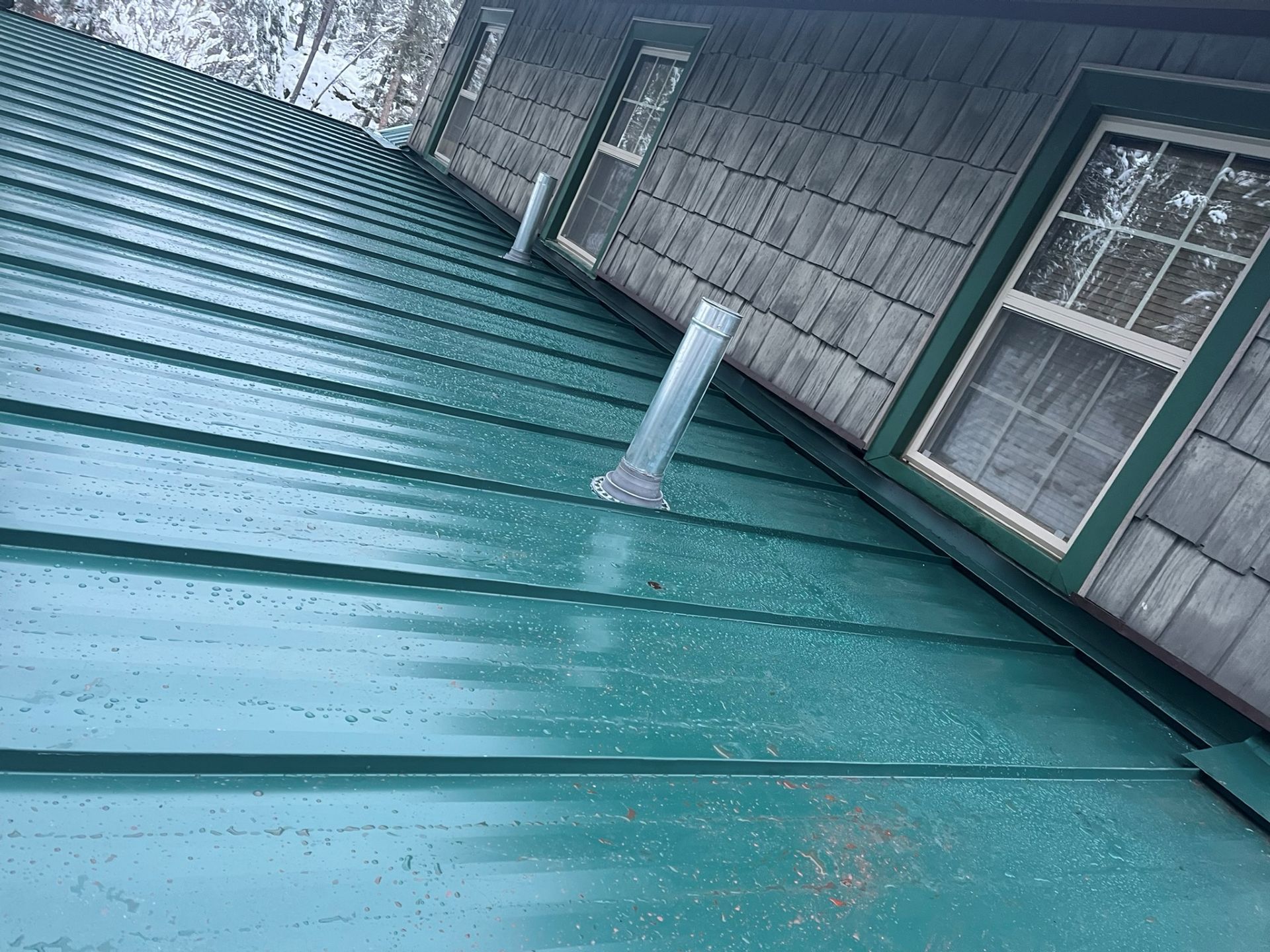 Green metal roof with water runoff pipe beside a gray building and window