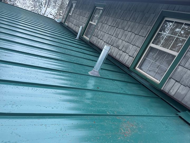 Tilted view of a teal metal roof with a white vent pipe beside a windowed building wall