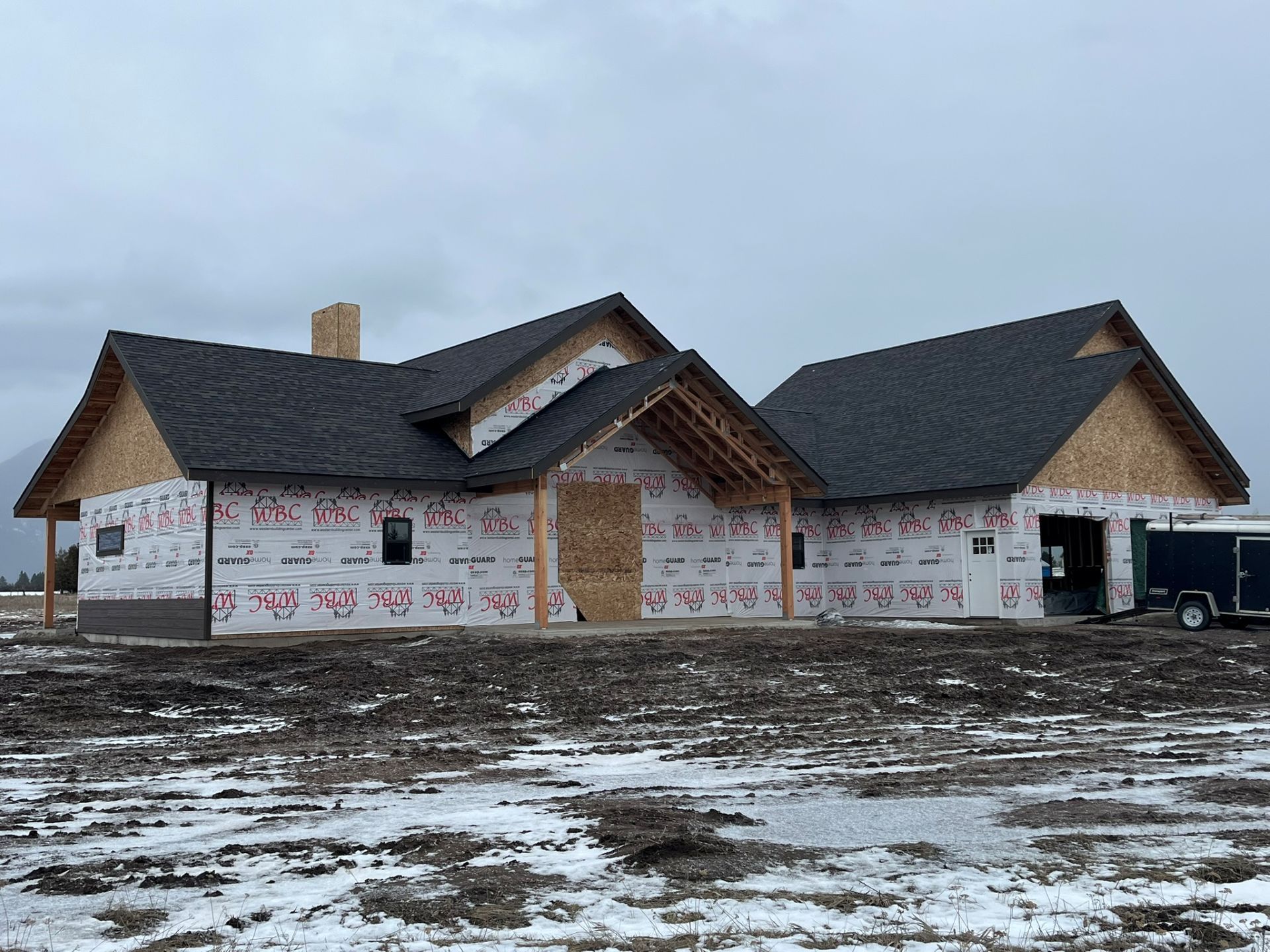 Large unfinished house with dark roof, wrapped in construction paper, on a snowy lot under a cloudy sky
