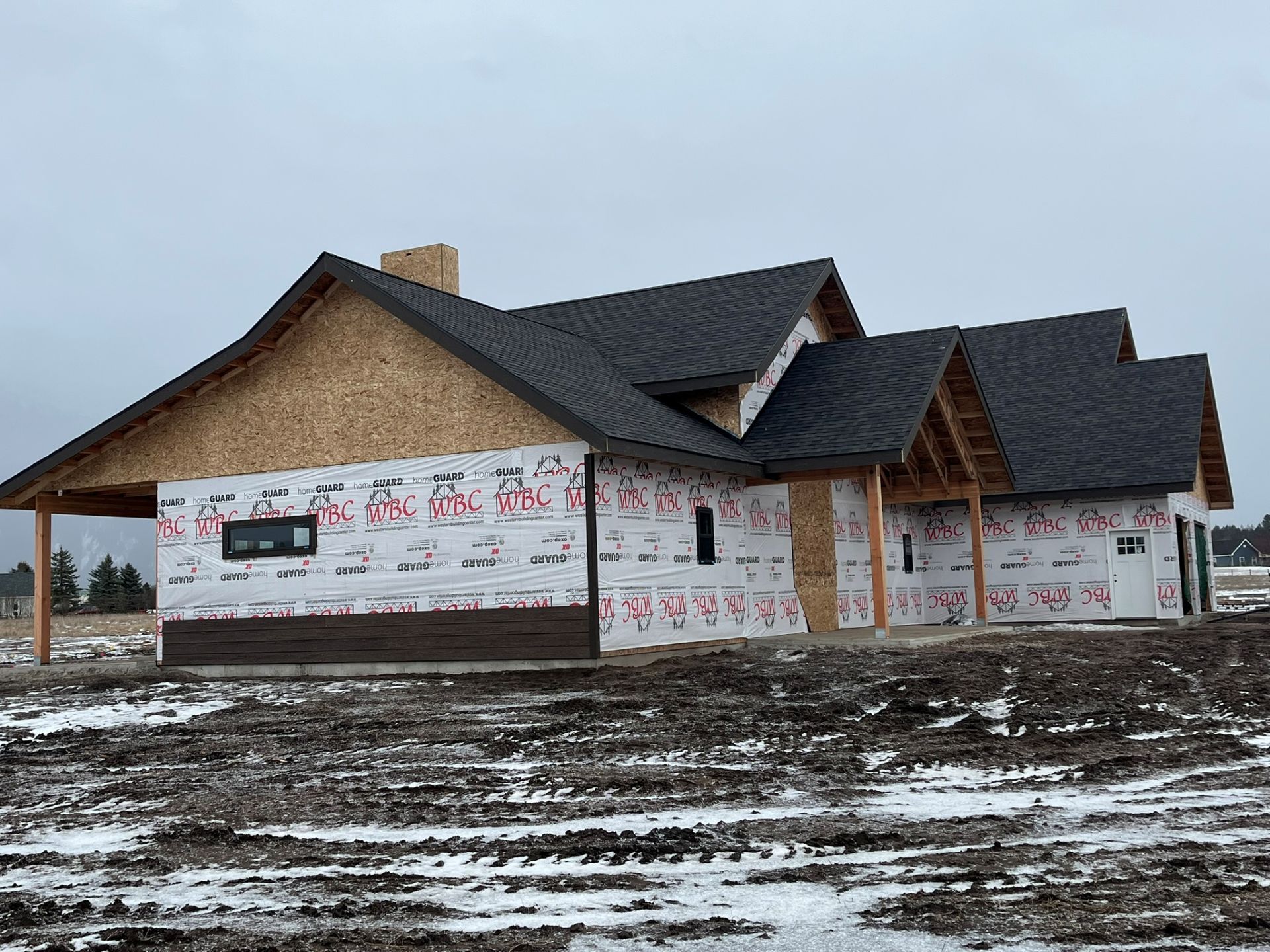Partially built house with dark roof and white wrap in a snowy, muddy lot under a gray sky