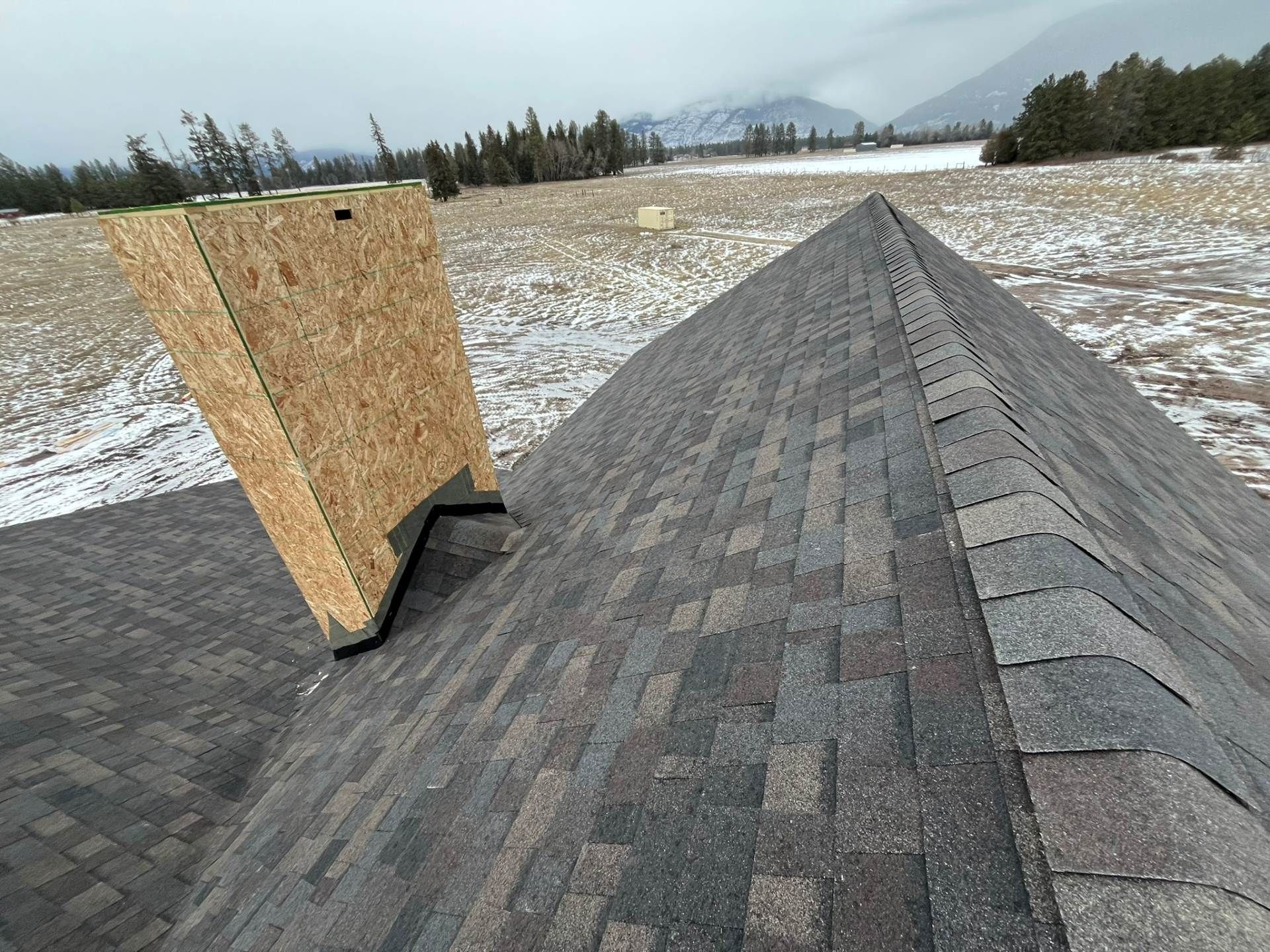 Snow-dusted rooftop with a plywood chimney cover and a long ridge of gray shingles under cloudy skies