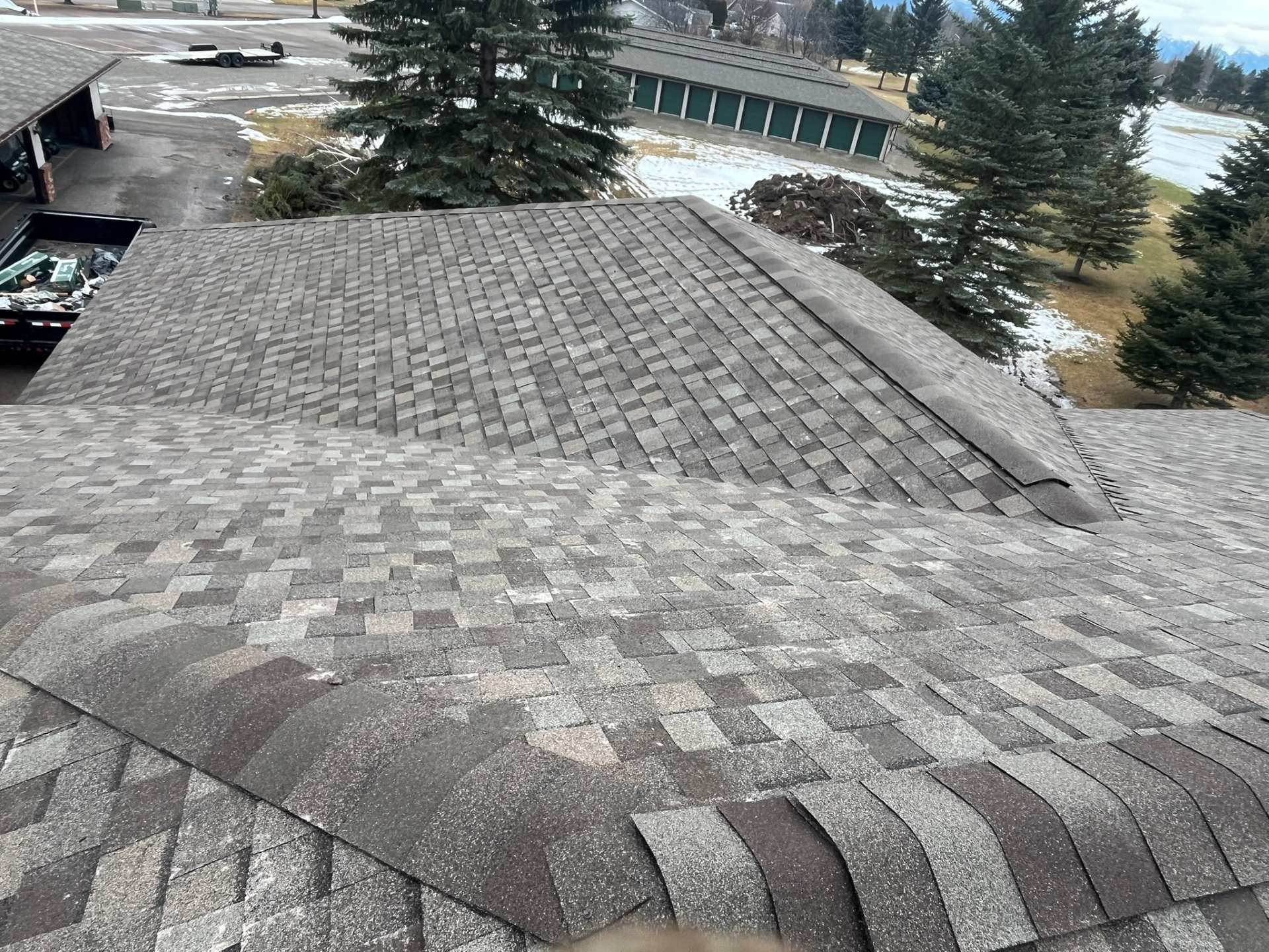 Snow-dusted shingled roof with tall trees and a curved building in the background