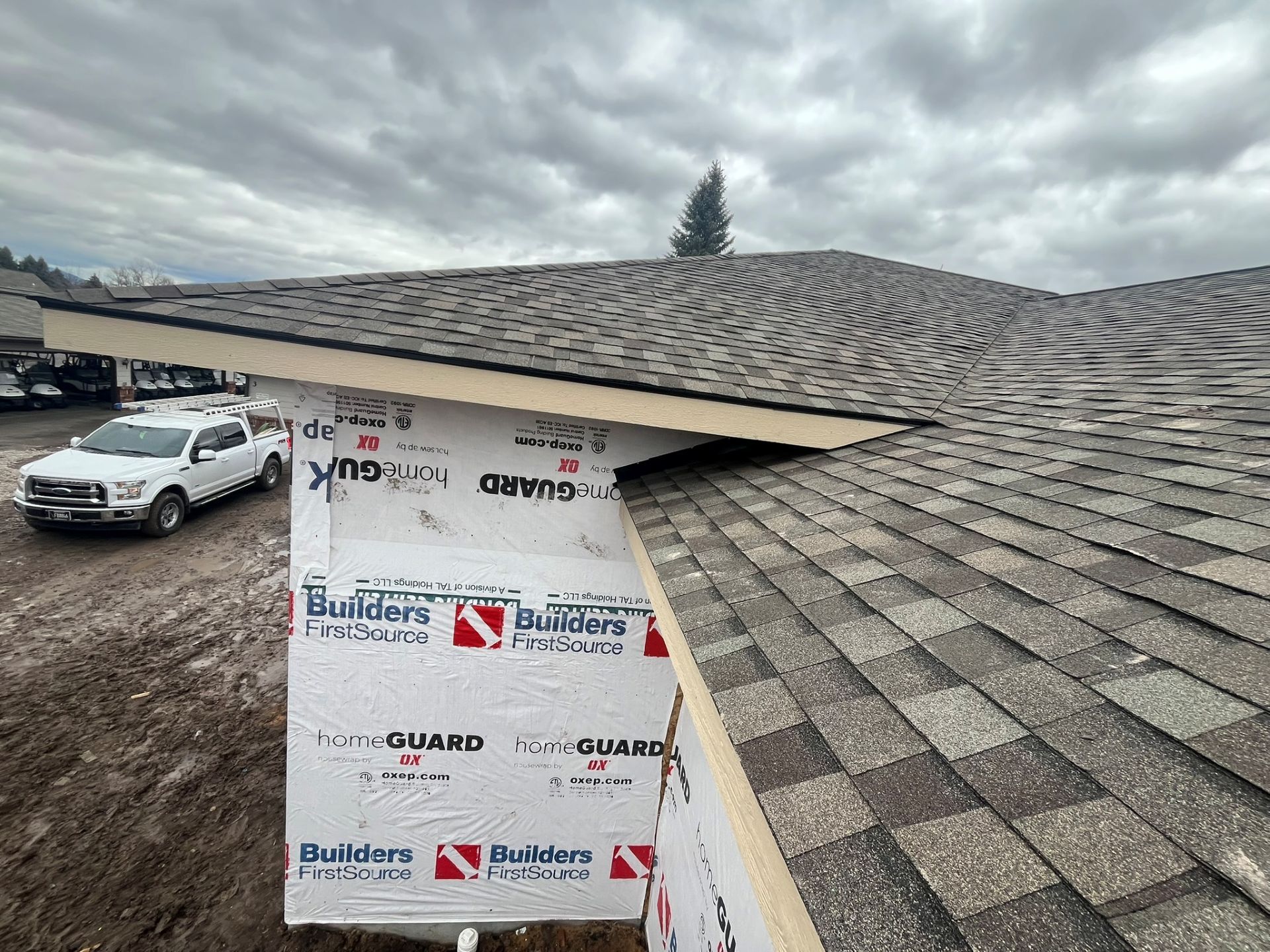 Damaged shingle roof with a hole exposing underlayment and roof deck, parked car below under cloudy sky