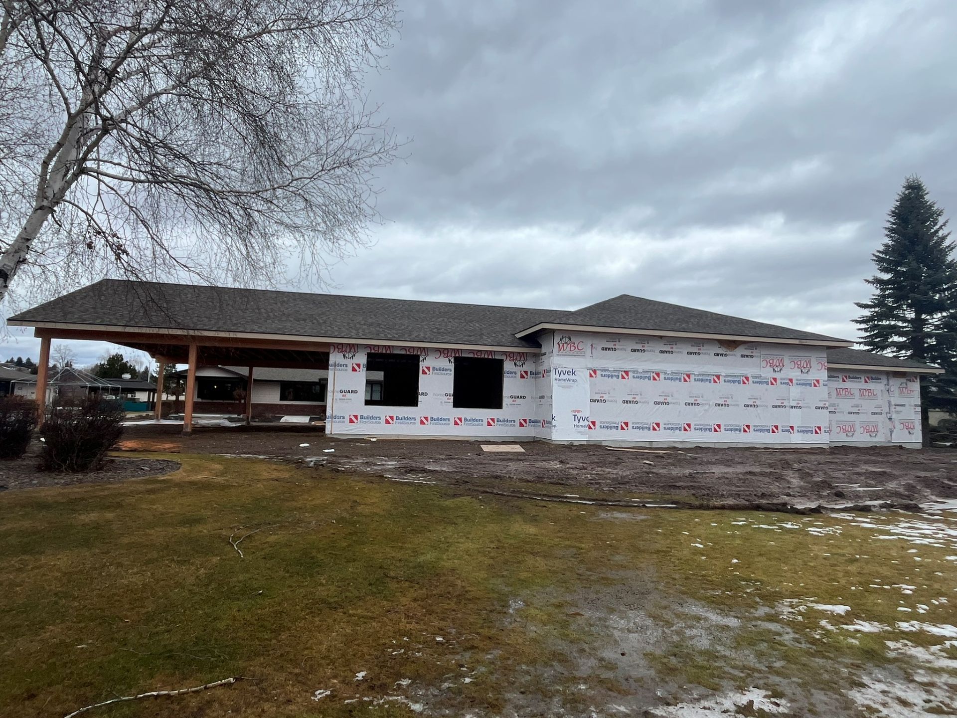 Single-story house under construction with white siding, bare yard, and overcast sky