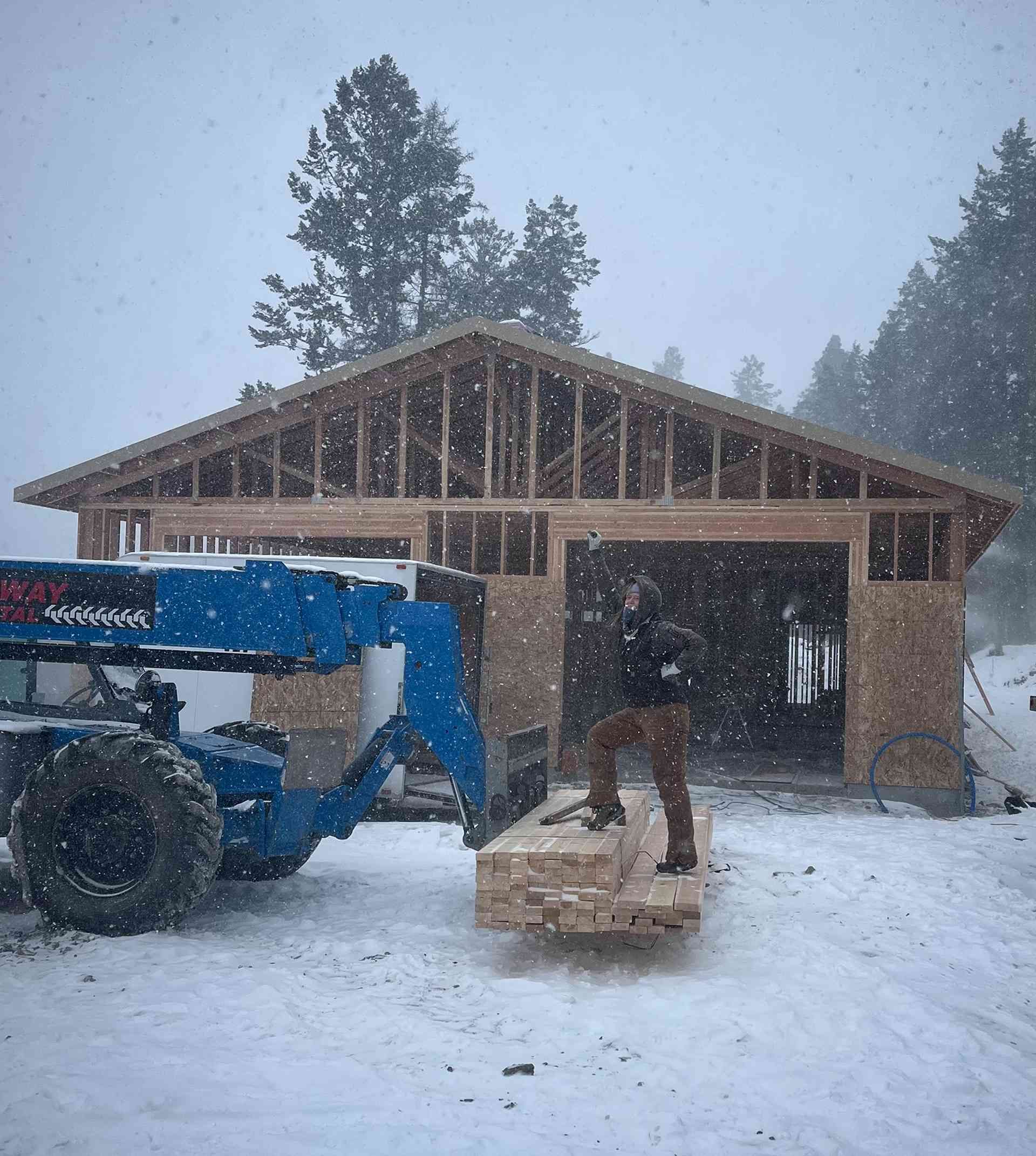 Blue tractor and worker in snowy construction site beside a partially built wooden garage.